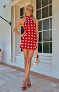 A full-length lifestyle shot of our model wearing a short red dress with large white polka dots. This dress features a high-neck halter neckline and a sleeveless form-fitting silhouette.