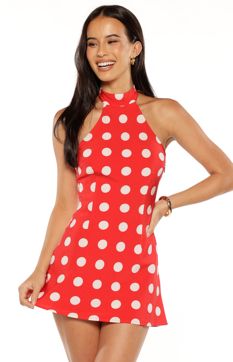 A front-facing studio shot from the neck down of our model wearing a short red dress adorned with large white polka dots. This mini dress features a sleeveless form-fitting silhouette with a high-neck halter design.